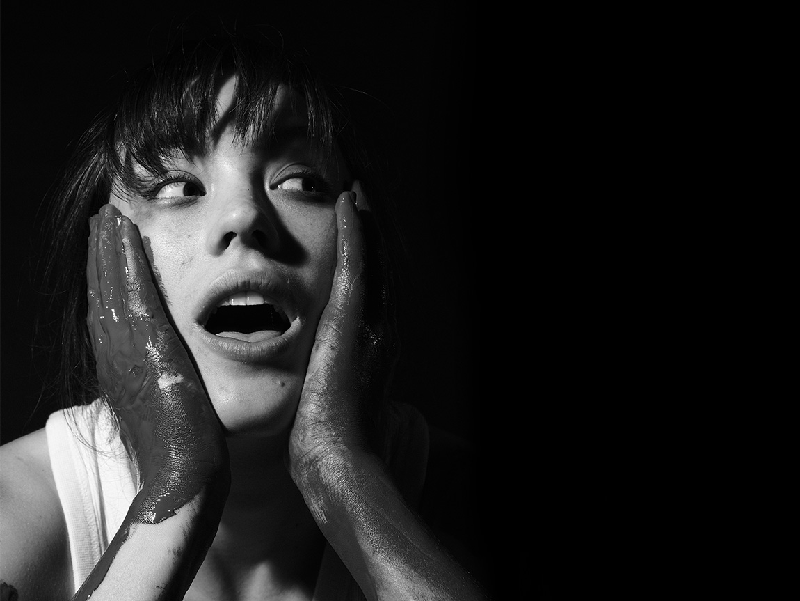 black-and-white photo of Katrina Binks. She has her hands to the sides of her face, and her hands are covered in something that could be paint. She is looking off to one side with her mouth open to suggest surprise.